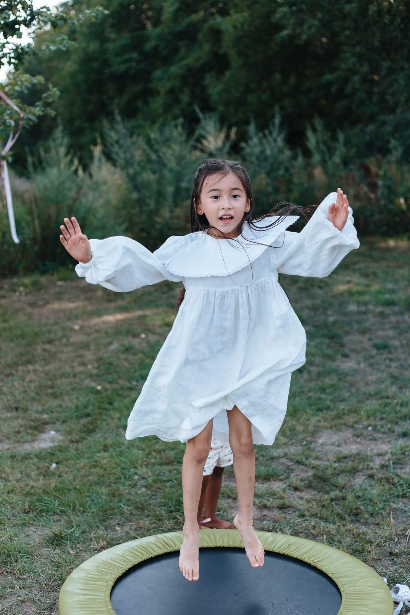 Happy girl in a white dress jumping on a trampoline in a green backyard.